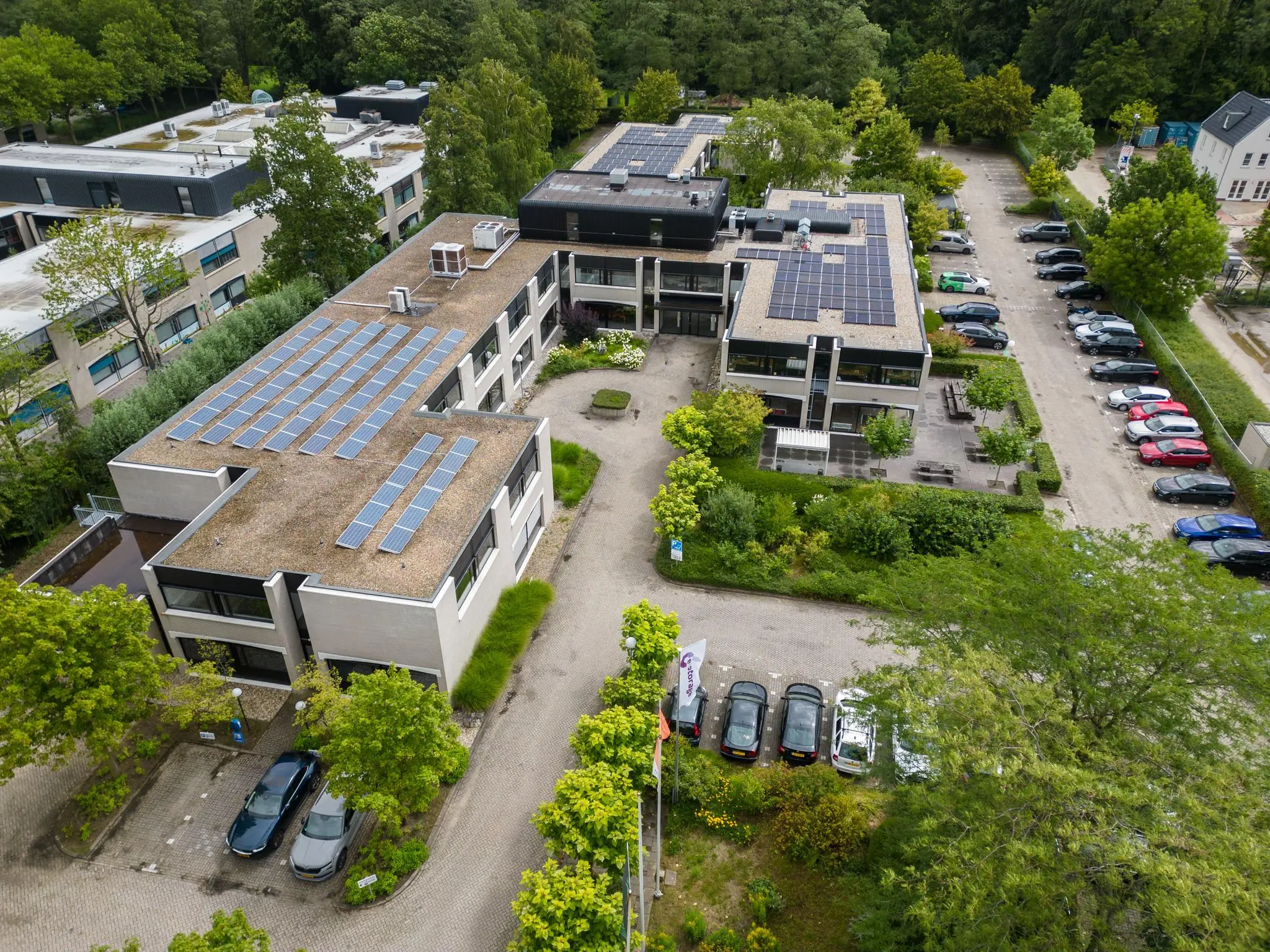 Luchtfoto van het gebouw aan de Burgemeester Verderlaan met zonnepanelen op het dak en omliggende parkeerplaatsen.